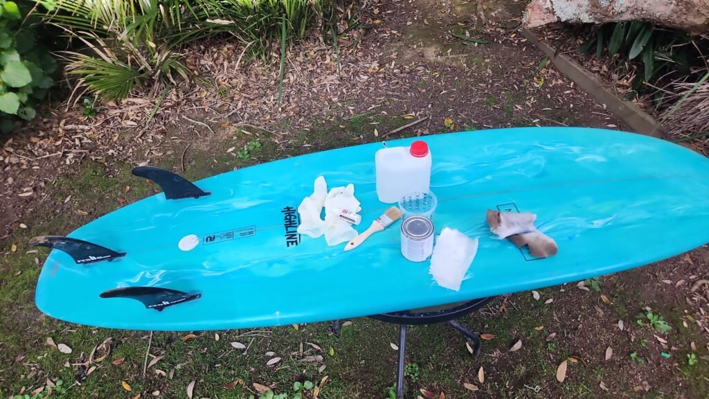 Planche de surf posée à plat avec le kit de réparation complet : résine, catalyseur, gants, pinceau et tissu de fibre de verre